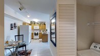 This single-floor residence offers a thoughtfully arranged layout that maximizes functionality and comfort. The kitchen serves as a central highlight, featuring warm honey-toned wood cabinetry that extends to the upper walls, topped with decorative greenery that adds a welcoming touch. White appliances — including a refrigerator, electric range, microwave, and dishwasher — are neatly integrated throughout the space. A central kitchen island with a wood-toned finish provides additional prep and storage space, while the sink is conveniently positioned nearby. Light-toned ceramic tile flooring spans the kitchen and adjoining areas, offering both durability and a clean, cohesive aesthetic. Track lighting on the ceiling ensures the space is well-lit and practical. A louvered shutter door adds architectural character and a classic design element near the entryway, alongside a framed mirror that enhances the sense of openness. The overall ambiance is bright, tidy, and functional, with decorative accents lending a lived-in warmth. The single-floor plan ensures easy navigation throughout, making this a practical and inviting space suited for everyday living.