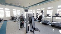 This fitness center is designed to support a comprehensive workout experience within a single open-plan floor. The space is filled with an array of professional-grade equipment, including strength machines from brands such as FreeMotion and Instinct, a dumbbell rack with multiple weight selections, elliptical trainers, a stair climber, and treadmills. The layout is thoughtfully organized, separating cardio and strength zones to allow for efficient use of the space. A wall-mounted television is positioned for visibility across the floor.

Architecturally, the room features a tray ceiling accented with a teal border, recessed lighting, and HVAC vents integrated cleanly into the ceiling. Large windows line the walls, allowing abundant natural light to flood the interior and offering views of the surrounding outdoor environment, including what appears to be mature landscaping. The light gray carpet provides a neutral, clean aesthetic underfoot. The overall ambiance is bright and modern, creating an inviting environment conducive to focused exercise. The combination of ample natural light, quality equipment, and refined architectural details gives the fitness center a polished, resort-style character.