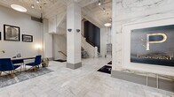 This distinguished lobby makes an immediate impression with its expansive, floor-to-ceiling marble wall cladding, displaying dramatic white and grey veining that lends the space a sense of timeless grandeur. The polished marble tile flooring continues the sophisticated aesthetic underfoot, laid in a classic brick pattern that guides visitors through the space. Structural columns rise prominently through the open floor plan, complementing the exposed ceiling with its industrial-style beams and track lighting that illuminate the area with a warm, focused glow. A sweeping marble staircase with elegant brass railings serves as a central architectural focal point, anchoring the space with a sense of historic prestige. Adjacent to the staircase, a bank of three elevators is framed by richly veined marble surrounds, each marked with polished metallic finishes. The Provident-branded reception area features a sleek dark signage panel with a gold logo, accompanied by a functional desk area dressed with blue velvet chairs, brass-accented furnishings, and a curated gallery of vintage black-and-white architectural photographs. The lobby's generous ceiling height amplifies the sense of openness, while glass entry doors at the front invite abundant natural light, completing this refined and welcoming arrival experience.