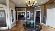 Located at 10925 Park Ave S in Tacoma, Washington, this single-floor community clubhouse offers a well-appointed gathering space designed for comfort and convenience. The entry foyer sets an inviting tone with warm earth-toned walls, large-format tile flooring in a classic checkerboard pattern, and an elegant chandelier overhead. A decorative round table anchors the space, while interior windows provide sightlines into adjacent rooms, including the fitness center and lounge beyond.

The main lounge is generously proportioned, featuring vaulted ceilings with a ceiling fan, recessed lighting, and an arched window that draws in abundant natural light. A tile-surround fireplace with a dark mantle serves as the focal point, complemented by a wall-mounted television. Comfortable leather seating and French doors opening to the outdoors make this an ideal space for relaxation or social gatherings. A kitchenette with cabinetry, a microwave, and a bar-height counter is conveniently positioned nearby.

A secondary seating area offers additional lounge space with high ceilings, warm accent walls, framed artwork, and a classic armoire. The dedicated fitness center is fully equipped with cable machines, free weights, kettlebells, resistance equipment, and cardio machines, all set on durable flooring with mirrored walls and direct outdoor access via French doors.