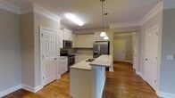 This single-floor residence offers a thoughtfully designed layout centered around a standout kitchen. The kitchen is the heart of the home, showcasing crisp white cabinetry with crown molding details that extend to the ceiling, adding a sense of height and elegance. Light granite countertops provide ample prep space and are complemented by a central island with a built-in sink, ideal for both cooking and casual entertaining. Pendant lighting hangs gracefully above the island, adding a warm, focused glow to the space. Stainless steel appliances, including a French door refrigerator with bottom freezer, a range with electric cooktop, and an over-the-range microwave, lend a modern and functional touch. Warm hardwood flooring flows throughout, tying the kitchen seamlessly to the adjoining living areas and hallway. Crown molding accents the ceilings, reinforcing the home's classic, refined character. Natural light filters through the space, enhancing the bright and open atmosphere. A hallway visible beyond the kitchen leads to additional rooms, suggesting a well-organized and practical floor plan. Overall, this home presents a clean, move-in-ready aesthetic with quality finishes throughout.