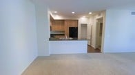 This single-floor residence showcases an open-concept design that seamlessly integrates the kitchen and living area. The kitchen is outfitted with warm honey-toned wood cabinetry providing ample storage, dark granite countertops, and black appliances including a refrigerator and microwave. A peninsula with a built-in sink and matching granite surface extends into the space, creating a natural division between the kitchen and the carpeted living area while offering additional workspace and casual seating opportunities. Recessed lighting throughout the ceiling ensures consistent, even illumination across both spaces. The living area features plush, light-toned carpeting, contributing to a comfortable and inviting atmosphere. Hardwood-style flooring is visible in the hallway beyond, which leads to additional rooms, including what appears to be a walk-in closet and a bathroom. The white walls and neutral color palette throughout create a clean, versatile backdrop. The layout is efficient and well-organized, with a hallway connecting the main living spaces to the private areas of the unit, offering a logical and practical flow throughout.