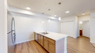 Located in Spokane, WA, this one-bedroom, one-bathroom residence offers a thoughtfully designed layout across a single floor. The open-concept kitchen is a standout feature, showcasing warm wood cabinetry, white countertops, stainless steel appliances including a refrigerator and range, a built-in dishwasher, and a large peninsula with an integrated sink and pendant lighting overhead. The kitchen flows naturally into the living area, which features light wood-style flooring near the entry and plush carpet in the main lounge space. The living room benefits from a glass-panel entry door and surrounding windows that invite natural light and outdoor views, creating a bright and welcoming atmosphere. The bedroom is a comfortable size, carpeted, and includes a window with blinds that provides additional natural light. An in-unit washer and dryer, visible in the overhead view, add everyday convenience. The bathroom is equipped with a full soaking tub and shower combo, complete with stainless grab bars and a handheld showerhead, reflecting accessible design. Clean white walls and consistent finishes throughout give the space a cohesive, modern feel.
