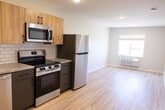 Unit C, #305 at Faribault offers 470 square feet of thoughtfully designed living space across one floor, comprising a combined kitchen/living room, one bedroom, and one bathroom. The unit features light wood-tone luxury vinyl plank flooring throughout, complemented by neutral, warm-toned walls and clean white trim. The kitchen is a standout feature, equipped with stainless steel appliances including a refrigerator, dishwasher, electric range, and built-in microwave. Two-toned cabinetry pairs natural wood upper cabinets with navy blue lower cabinets, accented by a classic white subway tile backsplash and light countertops. The open layout flows into a bright living area with a wall-mounted thermostat and exterior door access. The bedroom measures 110 square feet and includes a closet with a hanging rod and direct access to the main living space. It is equipped with a wall-mounted HVAC unit and a door leading to an outdoor area with exterior views. The bathroom features a full tub/shower combo, a wood-toned vanity with a white countertop, a round mirror, and a recessed ceiling light. This compact, modern unit provides efficient and functional living with contemporary finishes throughout.