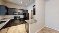 This single-floor residence offers a thoughtfully designed layout with modern finishes throughout. The kitchen serves as a functional centerpiece, outfitted with dark espresso cabinetry, granite countertops, a full suite of black appliances including a range, microwave, and refrigerator, and a peninsula that opens toward the living area. Track lighting illuminates the kitchen workspace, while light hardwood-style flooring flows seamlessly through the main areas. The open-concept living and dining space is bright and inviting, enhanced by a stylish orb chandelier and an abundance of natural light. The dining area features a window with blinds and the same warm chandelier detail, creating a welcoming atmosphere. Bedrooms are generously sized with clean neutral walls and hardwood-style flooring, with at least one bedroom offering direct access to a private bathroom. The bathrooms feature dark wood vanities, marble-style countertops, large mirrors, and hardwood-style tile flooring. A dedicated in-unit laundry room houses a full-size washer and dryer beneath a vaulted ceiling with overhead lighting and a built-in shelf. A walk-in pantry or storage closet with built-in shelving adds further practicality to this well-rounded residence.