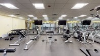 This fitness center is designed to accommodate a full range of workout needs within a single, open-plan space. The layout is organized to allow clear separation between cardio and strength-training zones, maximizing usability and flow. The flooring throughout consists of durable speckled rubber, a practical and slip-resistant surface well-suited for high-traffic exercise environments.

The cardio section is stocked with multiple treadmills, upright and recumbent stationary bikes, and elliptical machines, including Matrix-branded equipment. Two large flat-screen televisions are mounted on the mirrored walls, providing entertainment during workouts. Full-length mirrors line the walls, visually expanding the space and allowing users to monitor their form.

The strength-training area includes adjustable weight benches, cable machines, free weights, and multi-function cable stations. A glass partition with frosted decorative detailing separates a dedicated stretching or auxiliary workout area, offering a quieter zone within the overall space.

A drop ceiling with recessed panel lighting provides consistent, bright illumination throughout. Bold yellow accent bands run along the upper walls, adding a vibrant, energetic aesthetic that complements the functional design of the facility.
