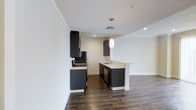 This single-floor space showcases a contemporary design aesthetic centered around a thoughtfully appointed kitchen. Dark, flat-front cabinetry creates a striking contrast against the crisp white walls and light-toned countertops, giving the space a clean, modern feel. Stainless steel appliances, including a refrigerator and a gas range, provide a professional-grade cooking environment. A kitchen peninsula extends into the open area, offering additional prep and seating space, and is accented by a sleek pendant light that adds a warm focal point to the room. Recessed lighting throughout ensures the space is well-lit and functional. Rich, dark hardwood flooring runs the length of the kitchen and into the adjoining area, grounding the space with warmth and texture. The layout is open and efficient, with upper and lower cabinetry providing ample storage on both sides of the kitchen corridor. The white walls and ceilings amplify the sense of openness, while the minimalist design choices keep the overall ambiance clean and uncluttered. This space is well-suited for those who appreciate modern design paired with practical, everyday functionality.