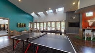 This single-floor community amenity space is designed with a bold, eclectic aesthetic that blends modern and rustic elements throughout. The entry foyer sets an immediate tone with a houndstooth-patterned accent wall, polished concrete floors, and statement signage reading "CA Wall – Eat Em Up," establishing a spirited, personality-driven atmosphere.

The main gathering area soars with high ceilings punctuated by multiple skylights, flooding the space with abundant natural light. A wall of floor-to-ceiling glass doors opens toward an outdoor pool area, seamlessly blending interior and exterior environments. This central zone is anchored by a full-size pool table and a ping-pong table, making it a dedicated game and recreation hub.

Adjacent to the game area, a lounge zone features a bold teal accent wall mounted with a large flat-screen TV, complemented by plush seating arranged around a round coffee table on a textured area rug. The space transitions naturally into a bar and lounge nook distinguished by rich terracotta-red patterned wallpaper, a horse portrait, warm sconce lighting, a wet bar with countertop appliances, and a communal high-top table set on artificial turf. Additional mounted televisions, decorative deer head mounts, and curated artwork complete the cohesive, ranch-inspired modern design.