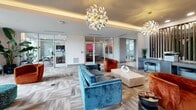 Located at 4950 Lunar Ln in Kissimmee, Florida, this impressive single-floor community space spans a generously sized footprint designed to foster comfort and social connection. The layout flows seamlessly from a bright, open entry corridor — finished with large-format light tile flooring and a dramatic coffered ceiling accented by warm LED cove lighting — into a series of distinct yet interconnected lounge areas.

The main living area serves as the centerpiece, featuring sculptural chandeliers with cascading metallic ribbons that draw the eye upward. Seating arrangements of teal velvet sofas, burnt orange armchairs, and patterned ottomans are thoughtfully positioned atop layered area rugs, creating intimate conversation zones within the larger open space. Floor-to-ceiling glazed walls and expansive windows flood the interior with natural light and offer views of the surrounding landscaped grounds.

A bar-height counter area with pendant lighting and upholstered stools provides an additional social hub, while glass-partitioned rooms along the perimeter offer quieter, semi-private spaces. Lush tropical plants are positioned throughout, reinforcing a fresh, resort-inspired atmosphere. Curated artwork, including a striking circular brushstroke piece and a living green wall near the entrance corridor, add distinctive artistic character to the space.