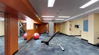 This fitness center is designed to provide a well-rounded workout environment within a single-floor layout. The space features bold burnt-orange accent walls paired with soft beige tones, creating an energetic yet functional atmosphere. Recessed rectangular fluorescent panels illuminate the room evenly across a dropped ceiling, and patterned blue-gray carpet tiles cover the entire floor area.

Cardio equipment includes a treadmill and an upright stationary bike, positioned near small windows that allow modest natural light into the space. A wall-mounted television is situated nearby for entertainment during workouts. A multi-station cable machine anchors the strength-training area, accompanied by an adjustable weight bench suitable for a variety of exercises. A three-tier dumbbell rack lines one wall, offering a range of free weights, while stability balls are available for core and flexibility training.

Large mirrors are built into one wall section, framed by the orange accent columns, visually expanding the space and aiding in form correction. Safety equipment, including a fire extinguisher and emergency signage, is clearly mounted on the walls. The room is accessible via a standard door and maintains a clean, organized layout conducive to efficient individual workouts.