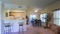 This single-story residence is designed with an open-concept layout that promotes a seamless flow between the kitchen, dining, and living areas. The kitchen features light maple cabinetry, a built-in microwave, and a raised breakfast bar with barstool seating — ideal for casual dining or entertaining. The pass-through design connects the kitchen directly to the main living space, enhancing the sense of openness throughout.

The dining area accommodates a full-sized dark wood table and chair set, positioned centrally within the shared living space. A ceiling fan with an integrated light fixture provides both comfort and ambient lighting overhead. Vaulted ceilings in the kitchen area add vertical dimension and a sense of spaciousness to the interior.

Natural light pours in generously through large sliding glass doors that lead to an outdoor patio or balcony, offering views of exterior greenery. Additional windows dressed with neutral curtains and blinds further brighten the space. The soft, neutral-toned carpeting throughout the living and dining areas contributes to the warm and inviting ambiance. Decorative indoor plants add a natural, refreshing element to the overall aesthetic. The space is well-suited for comfortable everyday living.
