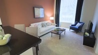 This single-floor space showcases a thoughtfully designed living area defined by warm terracotta-toned accent walls that create an inviting, cozy atmosphere. The living room centers around a classic white sofa adorned with decorative throw pillows in soft floral and botanical patterns, complemented by a dark-stained wooden coffee table that provides a stylish contrast to the light-toned carpeted flooring. A blue accent chair positioned near the large window adds a pop of color and visual interest to the room.

Natural light flows generously through a large window dressed with floor-length charcoal drapes, brightening the space while offering privacy control. A floor lamp beside the sofa provides warm supplemental lighting, enhancing the room's welcoming ambiance. Framed artwork on the accent wall adds a curated, gallery-like touch to the space.

A dark-surfaced counter or bar area is visible at the edge of the living room, suggesting a seamless transition into an adjacent dining or kitchen area. The neutral carpeting throughout ties the space together, contributing to an overall atmosphere that is both comfortable and refined.