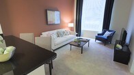 This single-floor space showcases a thoughtfully designed living area defined by warm terracotta-toned accent walls that create an inviting, cozy atmosphere. The living room centers around a classic white sofa adorned with decorative throw pillows in soft floral and botanical patterns, complemented by a dark-stained wooden coffee table that provides a stylish contrast to the light-toned carpeted flooring. A blue accent chair positioned near the large window adds a pop of color and visual interest to the room.

Natural light flows generously through a large window dressed with floor-length charcoal drapes, brightening the space while offering privacy control. A floor lamp beside the sofa provides warm supplemental lighting, enhancing the room's welcoming ambiance. Framed artwork on the accent wall adds a curated, gallery-like touch to the space.

A dark-surfaced counter or bar area is visible at the edge of the living room, suggesting a seamless transition into an adjacent dining or kitchen area. The neutral carpeting throughout ties the space together, contributing to an overall atmosphere that is both comfortable and refined.