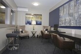 This single-floor professional space is thoughtfully designed to balance comfort and functionality. The interior features a consistent color scheme of cool blues and warm neutrals, with accent walls in deep navy blue adding depth and visual interest throughout. Durable, patterned carpet tiles in shades of grey and blue run throughout the space, providing both practicality and style.

A cozy lounge area anchors one section of the space, furnished with a pair of plush wingback chairs in a light cream upholstery, complemented by accent pillows in navy and grey. A glass-top side table with a sleek metallic base holds a table lamp, and a striking abstract artwork in gold, blue, and grey tones hangs above. A wall-mounted mini-split air conditioning unit ensures year-round climate comfort.

Adjacent to the lounge, a functional work area includes a dark wood desk with hairpin legs, seating for multiple users, and a desktop computer setup — ideal for focused tasks. A credenza with a wood tile finish houses a printer, adding to the workspace's practicality. A small glass-top bistro table with modern chairs offers additional flexible seating.

A glass-panel entry door provides a clear sightline to the surrounding corridor, while inset pass-through windows in the walls suggest a reception or service-oriented function. The overall ambiance is professional yet inviting, making it well-suited for an office, business center, or client-facing environment.