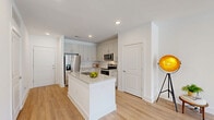 Located at 3025 Woodward Crossing Boulevard in Buford, GA, this thoughtfully designed single-floor residence offers a bright, modern living experience. The open-concept layout seamlessly connects the kitchen and living room, creating an inviting and functional common area bathed in natural light from large sliding glass doors and windows.

The kitchen is a standout feature, showcasing white shaker cabinetry extending to the ceiling, light granite countertops, a herringbone tile backsplash, and a full suite of stainless steel appliances including a refrigerator with ice maker, electric range, over-the-range microwave, and dishwasher. A central island with a built-in sink and additional granite countertop space provides both a prep area and casual dining option. Light wood-style flooring runs throughout the main living areas, enhancing the clean, contemporary aesthetic.

The spacious primary bedroom features plush carpeting, a ceiling fan, and a window that brings in natural light. The living area also opens to the outdoors via a sliding glass door. A dedicated in-unit laundry room houses a full-size washer and dryer with overhead wire shelving for added storage. The unit's crisp white walls and recessed lighting throughout create a bright, airy atmosphere well-suited to modern living.