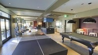 Located at 444 E. 19th Ave. in Denver, Colorado, this single-floor amenity space is a thoughtfully designed communal lounge that blends entertainment, socializing, and relaxation into one cohesive environment. The open-concept layout is anchored by light hardwood flooring and a modern suspended ceiling with recessed lighting, creating a bright and welcoming atmosphere throughout.

The game area is a standout feature, offering both a ping-pong table and a full-size billiards table beneath a sleek pendant light — ideal for casual recreation and social gatherings. Adjacent to the game area is a well-appointed wet bar, complete with a dark granite island, bar-height seating with contemporary blue stools, and a decorative tile backsplash that adds a bold visual accent. The bar area is equipped with ample counter space and cabinetry, making it well-suited for entertaining.

Beyond the bar, the lounge transitions into a comfortable seating area with upholstered furniture arranged around a fireplace, providing a warm and inviting retreat. Structural beams and varied ceiling heights lend architectural interest to the space, while the open sightlines maintain a sense of connectivity throughout. The overall design aesthetic is contemporary and polished, reflecting Denver's modern urban style.