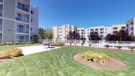 This residential complex presents a welcoming exterior defined by its modern, multi-story buildings clad in neutral beige and tan siding with crisp white trim accents. The buildings are arranged to frame a generously sized central courtyard that serves as the heart of the community. Lush, manicured lawns stretch across the shared outdoor space, complemented by curved garden beds filled with a variety of ornamental shrubs and seasonal plantings in rich tones of red, green, and gold. Smooth concrete walkways wind through the landscaped grounds, connecting building entrances and providing easy access throughout the property. White railing fencing defines portions of the outdoor area, adding a clean, structured aesthetic to the open space. A patio area with seating offers residents a dedicated spot to gather and relax outdoors. Each building features private balconies with white railings, offering residents personal outdoor space with views of the surrounding grounds. Ground-level unit entrances are framed by covered entryways and surrounded by manicured plantings, enhancing curb appeal at every access point. The overall ambiance of the complex is bright, airy, and well-kept, with the open courtyard layout and abundant natural light creating an inviting atmosphere throughout the community.
