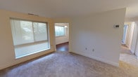 This single-floor residence offers a thoughtfully laid-out interior with a clean, neutral aesthetic throughout. The living area is generously sized, featuring wall-to-wall carpeting, soft neutral-toned walls, and large windows fitted with horizontal blinds that allow for ample natural light. An open passage connects the living space to an adjoining room with hardwood-style flooring, adding visual variety to the layout.

The galley-style kitchen is well-equipped with warm honey-toned wood cabinetry lining both sides, dark granite-patterned countertops, and white appliances including a gas range, dishwasher, and refrigerator. Overhead flush lighting illuminates the workspace efficiently, and the kitchen connects directly to the rest of the unit.

The bedrooms are carpeted and bright, each featuring windows with blinds for light control and privacy. One bedroom includes direct access to a spacious walk-in closet with built-in shelving, as well as an en suite bathroom. The bathrooms are identically appointed, each featuring an extended white vanity countertop, an oversized wall mirror with a globe light bar above, chrome fixtures, and a tiled shower/tub combination. The overall ambiance of the unit is clean, functional, and move-in ready.