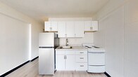 This efficiently designed studio offers a thoughtfully organized layout across a single floor, making smart use of every square foot. The open-concept living area is anchored by a large floor-to-ceiling window that floods the space with natural light and provides sweeping elevated views of the surrounding cityscape. Light wood-tone vinyl plank flooring runs throughout, complementing the bright, neutral color palette that gives the unit an airy, modern feel.

The kitchen is neatly integrated into the space and features white shaker-style cabinetry extending to the ceiling for generous storage, a stainless steel sink with a modern faucet, light marble-patterned countertops, a full-size refrigerator, and a white electric range. A textured accent wall adds subtle visual interest to the main living area.

A hallway connects the living space to the private areas of the unit, including a closet for storage and a full bathroom. The bathroom is clean and functional, featuring light wood-tone vinyl flooring, a white vanity with cabinet storage, a full soaker tub with an overhead shower, and brushed gold fixtures that add a refined touch. Wall-mounted sconce lighting in the hallway contributes to a warm, welcoming ambiance throughout the unit.