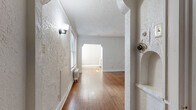 This single-floor space showcases a compelling mix of vintage character and functional updates. The entry corridor features distinctive textured walls, a wall sconce, and a decorative built-in niche, leading through an arched passageway into the main living areas. Warm hardwood floors flow throughout, anchoring each room with a sense of continuity and timeless appeal.

The living room is bright and inviting, benefiting from multiple windows dressed with blinds that allow generous natural light. Classic wainscoting panels line the walls, complemented by a cast-iron radiator that adds to the vintage aesthetic. Crown molding and a textured ceiling contribute additional architectural interest. An arched doorway connects the living space to adjoining rooms, reinforcing the period-inspired design language found throughout.

The kitchen has been thoughtfully updated with light grey shaker-style cabinetry, warm butcher block countertops, a textured tile backsplash, and stainless steel appliances including a gas range and refrigerator. The bedroom offers a spacious, open feel with hardwood floors, textured walls, and crown molding, accompanied by a generous walk-in closet with built-in shelving. The bathroom retains its classic white subway tile surround and a window above the tub, adding a traditional finishing touch to this characterful space.