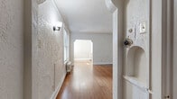 This single-floor space showcases a compelling mix of vintage character and functional updates. The entry corridor features distinctive textured walls, a wall sconce, and a decorative built-in niche, leading through an arched passageway into the main living areas. Warm hardwood floors flow throughout, anchoring each room with a sense of continuity and timeless appeal.

The living room is bright and inviting, benefiting from multiple windows dressed with blinds that allow generous natural light. Classic wainscoting panels line the walls, complemented by a cast-iron radiator that adds to the vintage aesthetic. Crown molding and a textured ceiling contribute additional architectural interest. An arched doorway connects the living space to adjoining rooms, reinforcing the period-inspired design language found throughout.

The kitchen has been thoughtfully updated with light grey shaker-style cabinetry, warm butcher block countertops, a textured tile backsplash, and stainless steel appliances including a gas range and refrigerator. The bedroom offers a spacious, open feel with hardwood floors, textured walls, and crown molding, accompanied by a generous walk-in closet with built-in shelving. The bathroom retains its classic white subway tile surround and a window above the tub, adding a traditional finishing touch to this characterful space.