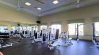 This fitness center offers a spacious, single-floor workout environment designed to support a full range of strength and conditioning exercises. The facility is outfitted with an extensive array of cable machines, weight stations, and resistance equipment, all arranged to allow comfortable movement throughout the space. Large arched windows along the walls flood the room with natural light, creating an open and energizing atmosphere, while a ceiling fan aids in air circulation during workouts. The high ceilings contribute to a sense of openness rarely found in typical gym settings. Durable rubber flooring covers the entire workout area, providing both safety and stability for users. Wall-mounted mirrors run along one side of the room, visually expanding the space and allowing users to monitor their form during exercise. Recessed and panel lighting in the drop ceiling ensures consistent illumination throughout the facility. A wall-mounted television is positioned for visibility across the floor. The warm, neutral-toned walls and clean layout give the space a professional yet welcoming ambiance, making it a functional and well-appointed environment for fitness enthusiasts of all levels.
