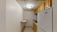 Located at 1144 Commonwealth Ave in Boston's Allston neighborhood, this single-floor unit offers a straightforward and functional layout across several interconnected rooms. The flooring plan, visible in the dollhouse view, reveals a series of open living and utility spaces connected by a corridor-style flow.

The kitchen is compact and efficiently arranged, featuring warm-toned wood cabinetry, a gas range, a stainless steel sink with under-counter storage, a refrigerator, and a built-in wall shelf that adds extra prep or storage space. A drop ceiling with recessed tile panels and a central light fixture keeps the area well-lit.

The living areas are finished with hardwood-style flooring that lends warmth throughout the unit. Drop-tile ceilings are consistent across rooms, and baseboard heating units run along the walls, providing efficient climate control. Small ground-level windows in one room allow for natural light to filter in, complementing the open, unfurnished space.

The bathroom is clean and practical, equipped with a white vanity with under-sink storage, a mirrored medicine cabinet above, and a full bathtub with a shower curtain enclosure. Light tile flooring ties the space together neatly.

Overall, this unit presents a no-frills, move-in-ready layout well-suited for practical urban living in a well-connected Boston location.