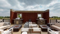 Located at 1050–1054 Station Blvd in Aurora, Illinois, this outdoor rooftop terrace is a well-appointed amenity space designed for communal gathering and al fresco entertaining. The terrace is anchored by two built-in stainless steel grilling stations, each set into stone-clad countertops and housed against a striking red brick wall featuring open window cutouts that frame views of the surrounding landscape. Flanking the central seating area are two rectangular fire pit tables filled with decorative glass rock, providing both ambiance and warmth during cooler evenings. The central lounge area features modern outdoor furniture, including a sectional sofa and accent chairs with neutral upholstered cushions, arranged to encourage conversation and relaxation. A wraparound walkway extends along the perimeter of the terrace, lined with potted evergreen topiaries and illuminated by classic lantern-style wall sconces. Black metal railings define the outer edges of the terrace, opening up to panoramic views of the Aurora skyline and surrounding greenery. The combination of functional grilling infrastructure, inviting fire features, and thoughtfully arranged seating makes this rooftop terrace a versatile outdoor retreat suited for both casual gatherings and larger social events.