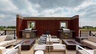 Located at 1050–1054 Station Blvd in Aurora, Illinois, this outdoor rooftop terrace is a well-appointed amenity space designed for communal gathering and al fresco entertaining. The terrace is anchored by two built-in stainless steel grilling stations, each set into stone-clad countertops and housed against a striking red brick wall featuring open window cutouts that frame views of the surrounding landscape. Flanking the central seating area are two rectangular fire pit tables filled with decorative glass rock, providing both ambiance and warmth during cooler evenings. The central lounge area features modern outdoor furniture, including a sectional sofa and accent chairs with neutral upholstered cushions, arranged to encourage conversation and relaxation. A wraparound walkway extends along the perimeter of the terrace, lined with potted evergreen topiaries and illuminated by classic lantern-style wall sconces. Black metal railings define the outer edges of the terrace, opening up to panoramic views of the Aurora skyline and surrounding greenery. The combination of functional grilling infrastructure, inviting fire features, and thoughtfully arranged seating makes this rooftop terrace a versatile outdoor retreat suited for both casual gatherings and larger social events.