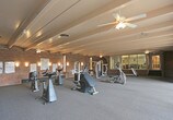 Located at 2201 Ambassador Rd NE in Albuquerque, New Mexico, this single-level fitness center offers a well-equipped workout environment defined by its distinctive architectural character. The space is framed by exposed brick walls and overhead beamed ceilings, giving it a sturdy, classic aesthetic that sets it apart from a typical gym setting.

The fitness floor is generously sized and carpeted throughout, providing a comfortable surface across multiple workout zones. The cardio area is equipped with stationary bikes, an elliptical trainer, and additional cardio machines, offering a solid range of low-impact exercise options. A Coca-Cola vending machine is conveniently positioned nearby. The strength training area features multi-station cable machines and weight benches, accommodating a variety of resistance exercises.

Large floor-to-ceiling windows along one wall flood the space with natural light and offer views of the outdoor surroundings, including mature trees and an exterior courtyard area. Ceiling fans help maintain airflow throughout the room. Wall-mounted instructional fitness charts and posted fitness center rules provide helpful guidance for users.

A glass partition separates a secondary room visible from the main floor, and a staircase near the entrance connects to adjacent areas. The overall ambiance is functional and straightforward, making this fitness center a practical and accessible facility for regular use.