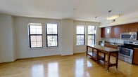This single-floor space offers a clean, modern aesthetic defined by an open-plan layout that connects the living area and kitchen in one cohesive environment. Gleaming hardwood floors run throughout, reflecting abundant natural light that pours in through multiple large, black-framed grid windows. These windows frame views of the surrounding neighborhood and lend the space a bright, airy quality throughout the day.

The living area is generously proportioned, offering a versatile, unobstructed floor plan well-suited for a variety of furniture arrangements. Neutral white walls and white ceilings complement the warm tones of the flooring, creating a balanced and inviting ambiance.

The kitchen transitions seamlessly from the open living space and features warm wood cabinetry, granite countertops, and stainless steel appliances. A freestanding wood and granite kitchen island with an open lower shelf adds both functional workspace and a stylish focal point to the room. Pendant lighting above the island provides targeted illumination, while a flush ceiling fixture serves the broader kitchen area.

Overall, the space combines practical design with a welcoming atmosphere, making it a well-lit, functional, and visually cohesive environment ideal for everyday living.