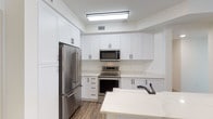 This single-floor space showcases a clean, modern aesthetic with thoughtful design choices throughout. The kitchen is a standout feature, outfitted with crisp white shaker-style cabinetry that extends from floor to ceiling, providing generous storage. Matte black hardware adds a sharp, contemporary contrast against the white finishes. Stainless steel appliances — including a French door refrigerator, electric range with oven, and over-the-range microwave — lend a professional, polished look to the space. Light quartz or stone countertops complement the cabinetry and contribute to the bright, airy atmosphere. A white kitchen island or peninsula offers additional workspace and casual seating potential. The sink is fitted with a sleek matte black faucet, consistent with the overall design language. Warm wood-look flooring grounds the space with texture and contrast, balancing the otherwise neutral palette. Recessed and flush-mounted overhead lighting ensures the kitchen is well-lit and functional. The overall ambiance is fresh, clean, and move-in ready, reflecting a contemporary design sensibility that prioritizes both style and practicality.