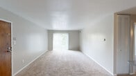 This single-floor unit is designed with a straightforward, functional layout that accommodates comfortable everyday living. The living area is notably open and bright, featuring neutral-toned carpet, light gray walls, and a large sliding glass door dressed with vertical blinds that fills the space with natural light and provides views of surrounding greenery outside.

The kitchen is equipped with warm oak cabinetry extending to the ceiling for ample storage, dark countertops, and wood-look flooring. Black appliances include a full-size refrigerator, dishwasher, and range, with a built-in microwave also present. The kitchen opens toward the living and dining areas, supporting an easy flow between spaces.

The bedrooms are carpeted and well-proportioned, each receiving natural light from windows. One bedroom features a wide wall of louvered bifold closet doors concealing a walk-in-style closet with wire shelving systems, providing generous storage capacity.

The bathroom includes a wood-vanity with a single sink, a large mirror with overhead lighting, and a full tub-shower combination with a tile surround. Wood-look flooring runs throughout the bathroom. A dedicated laundry room with wood-look flooring, washer/dryer hookups, and overhead wire shelving completes the unit.
