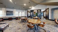 Located at 1000 Archive Cir in Oxford, Mississippi, this generously sized single-floor community amenity space offers a diverse range of social and recreational areas thoughtfully designed for comfort and engagement. The main lounge area impresses with soaring double-height ceilings, oversized windows that flood the room with natural light, and sophisticated furnishings including plush sofas, leather armchairs, and a live-edge wood coffee table, all anchored by a stylish area rug. An adjacent dining and social area features round wooden tables with curved leather chairs beneath statement pendant lighting, complemented by built-in booth seating along the walls. A dedicated coffee bar area stands out with a lush living green wall backdrop accented by a "Coffee, Please!" marquee sign, paired with a navy blue service counter and café-style seating. The entertainment lounge showcases a striking wood lattice ceiling, vertical slat wall paneling, a large wall-mounted screen, and an array of modern swivel lounge chairs. The game room delivers a full-size billiard table, a skee-ball arcade machine, and a pinball machine, with floor-to-ceiling glass walls offering direct views of an outdoor pool area. Warm hardwood-style flooring and soft blue accent walls create a cohesive, modern aesthetic throughout the entire space.