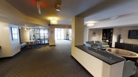 This single-floor space presents a well-organized common area designed to accommodate multiple functions within an open, connected layout. The main corridor runs through the center of the space, drawing the eye toward a bright, naturally lit entrance at the far end, flanked by a potted plant and a small accent table.

To the right, a built-in reception or service counter with a dark granite-style top anchors the space. Behind it, a comfortable lounge area features upholstered sofas and chairs arranged around a low table, with a tall bookcase lining the wall alongside framed artwork. Flush-mount, drum-style light fixtures provide warm ambient lighting throughout.

To the left, a windowed dining or meeting area is enclosed by dark-framed glass partitions, offering a semi-private setting furnished with tables and chairs. The large windows allow natural light to filter into this section of the space.

Underfoot, patterned commercial carpeting in a neutral tone covers the floor throughout, contributing to a quiet, functional ambiance. The warm yellow and beige wall tones, combined with recessed and surface-mounted lighting, create an inviting and practical environment suited for community or professional use.