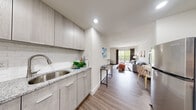 This single-floor studio unit presents a thoughtfully designed open-concept layout that seamlessly integrates the kitchen, living, and sleeping areas into one cohesive space. The kitchen runs along one side of the unit and is outfitted with light wood-grain cabinetry, granite countertops, a white subway tile backsplash, an undermount sink, and a full suite of stainless steel appliances including a refrigerator, electric range, and over-the-range microwave. Wood-look flooring extends throughout the entire unit, providing a warm and consistent aesthetic.

The combined living and sleeping area is generously proportioned, accommodating a full bed, dresser, seating area, and accent furniture without feeling cramped. A sliding glass door leads to a private balcony, allowing natural light to fill the space and offering views of surrounding greenery. A wall-mounted HVAC unit ensures individual climate control.

The bathroom is clean and functional, featuring a tiled walk-in shower with a curtain rod, a single-sink vanity, and wood-look flooring that complements the rest of the unit. Recessed lighting throughout maintains a bright, modern atmosphere across all areas of the space.