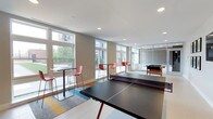 This single-floor recreational amenity space is thoughtfully designed to encourage social interaction and active play. The open layout accommodates a full-size ping pong table as a centerpiece, while a dedicated game area — partially enclosed by floor-to-ceiling glass partitions — houses a foosball table, offering a sense of separation without sacrificing the open feel of the room. High-top tables paired with modern coral-toned bar stools are arranged throughout, providing informal seating ideal for conversation or casual dining. Large windows along the exterior walls flood the space with natural light and offer views of the surrounding greenery, creating a bright and welcoming atmosphere. The interior design balances a clean, contemporary aesthetic with playful accents — colorful area rugs in yellow, blue, and gray add warmth and visual interest to the light tile flooring. Recessed ceiling lighting complements the abundant daylight, ensuring the space remains well-lit at all hours. The neutral wall tones and white ceilings keep the environment feeling open and airy, while the thoughtful arrangement of furnishings and games makes this an inviting destination for recreation and community gathering.