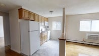 Located at 221 South Oak Avenue in Clifton Heights, Pennsylvania, this single-floor space offers a functional and well-organized layout designed for comfortable everyday living. The kitchen serves as a central feature, outfitted with warm-toned wood cabinetry that provides generous storage both above and below the countertops. White appliances, including a full-size refrigerator, electric range, and microwave, are neatly integrated into the layout. A window above the sink allows natural light to brighten the cooking area, enhancing the overall sense of openness. The kitchen flows directly into an adjoining living area, separated by a partial partition with a wood-finished countertop ledge that adds both visual definition and functional surface space. The living area features classic hardwood flooring, adding warmth and a timeless quality to the space. Light, neutral wall tones throughout contribute to an airy, cohesive atmosphere. A baseboard heating unit is visible along the wall, ensuring year-round comfort. The seamless transition between the kitchen and living areas creates an open, connected feel that makes the space practical and inviting.