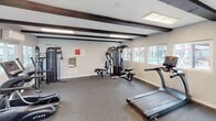 Located at 2960 E Jackson Ave in Anaheim, California, this fitness center offers a well-appointed, single-floor workout facility designed to support a well-rounded exercise routine. The space is characterized by its clean, light-colored walls and durable rubber flooring, providing a functional and comfortable environment for physical activity.

Distinctive dark exposed ceiling beams add an architectural contrast to the bright interior, while flush-mounted fluorescent lighting ensures the space is evenly and adequately illuminated throughout. A wall-mounted mini-split HVAC unit provides climate control for year-round comfort.

Multiple windows line the walls, allowing natural light to filter in and offering views of the surrounding outdoor amenity areas, including a pool and landscaped grounds. A dedicated entry door with direct outdoor access adds convenience for users.

The facility is equipped with a diverse selection of True-brand cardio machines, including treadmills, an elliptical trainer, and a rowing machine. A multi-station cable machine and functional trainer provide comprehensive strength training options. Free weights and additional accessories round out the equipment offerings. A built-in water fountain station is conveniently positioned along the wall, and posted fitness center rules and a welcome sign contribute to an organized and welcoming atmosphere.