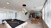 This well-appointed single-floor space is thoughtfully designed for entertaining and comfortable living. The open-concept layout flows seamlessly from one area to the next, creating a cohesive and inviting atmosphere throughout.

The entertainment zone anchors the space with a full-size pool table beneath a striking cascading pendant light fixture, whose clustered glass bulbs serve as a dramatic focal point. Light-toned luxury vinyl plank flooring runs throughout, lending warmth and continuity across the entire floor plan.

Adjacent to the billiards area, a relaxed seating nook features eclectic, modern furnishings including leather butterfly chairs and a natural wood stump side table, paired with a wall-mounted flat-screen TV for a laid-back lounge experience.

The open kitchen is outfitted with stainless steel appliances, a light-colored countertop bar with seating, and track lighting overhead — making it both functional and stylish. A dining area with a wood table and coordinating seating sits nearby, accented by an eye-catching piece of abstract wall art that adds a vibrant burst of color.

Recessed lighting throughout keeps the space bright and well-lit, while natural light filters in through windows near the entry. The overall ambiance is modern, casual, and well-suited for social events or communal use.