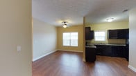 This single-story residence at Vineland Carriage Homes offers a practical and inviting layout designed for comfortable everyday living. The open-concept living area is finished with rich hardwood-style flooring that extends throughout, providing a warm and cohesive aesthetic. Warm yellow-toned walls brighten the interior, complemented by white baseboards that add a clean, classic contrast. A ceiling fan centered in the living space ensures year-round comfort while contributing to the room's casual, functional character. The textured ceiling adds subtle visual interest overhead.

Natural light flows generously into the living area through a large multi-pane window, creating an airy atmosphere. The kitchen is positioned adjacent to the living space, separated by a partial structural column that maintains an open feel between the two areas. Dark espresso-toned cabinetry provides bold contrast against the lighter walls, while granite-style countertops add a touch of refinement. A second window above the sink area brings additional natural light into the kitchen. The overall design balances practicality with comfort, making this a well-appointed single-floor home.