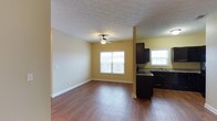This single-story residence at Vineland Carriage Homes offers a practical and inviting layout designed for comfortable everyday living. The open-concept living area is finished with rich hardwood-style flooring that extends throughout, providing a warm and cohesive aesthetic. Warm yellow-toned walls brighten the interior, complemented by white baseboards that add a clean, classic contrast. A ceiling fan centered in the living space ensures year-round comfort while contributing to the room's casual, functional character. The textured ceiling adds subtle visual interest overhead.

Natural light flows generously into the living area through a large multi-pane window, creating an airy atmosphere. The kitchen is positioned adjacent to the living space, separated by a partial structural column that maintains an open feel between the two areas. Dark espresso-toned cabinetry provides bold contrast against the lighter walls, while granite-style countertops add a touch of refinement. A second window above the sink area brings additional natural light into the kitchen. The overall design balances practicality with comfort, making this a well-appointed single-floor home.