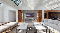 This thoughtfully designed single-floor space blends contemporary architecture with warm, natural materials to create an inviting and sophisticated environment. Soaring vaulted ceilings with exposed structural elements define the interior, while an abundance of skylights and large, floor-to-ceiling grid-pane windows flood every corner with natural light, creating an open and airy atmosphere throughout.

The central lounge area serves as the focal point of the space, anchored by a dramatic feature wall clad in textured gray stone tile. A wide linear fireplace sits beneath a wall-mounted television, flanked symmetrically by warm wood slatted panels and integrated wall sconces. Plush gray sofas and marble-topped coffee tables are arranged to encourage comfortable gathering and conversation.

An adjacent sunlit lounge corridor features large black-framed windows with sweeping exterior views, complemented by light tile flooring and a curated mix of upholstered armchairs and leather seating. The casual, flexible arrangement of furniture suits both social interaction and quiet relaxation.

The kitchen area is finished with sleek white cabinetry, a dark stone countertop, an undermount sink with a matte black faucet, and stainless steel appliances including refrigerator drawers — all accented by warm wood slatted wall panels that carry through the space's design language cohesively.