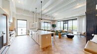 This single-floor space offers a seamlessly integrated open-concept layout encompassing a gourmet kitchen, dining area, and living room, all unified by striking hexagonal tile flooring and soaring white-painted beamed ceilings that amplify the sense of volume and light. The chef's kitchen is a standout feature, anchored by a generous island with a crisp white countertop and seating for multiple guests. Light wood cabinetry, glass-front upper cabinets, a professional-grade gas range, and a slate-blue refrigerator complete the well-appointed culinary space. Pendant lighting with elegant glass globes illuminates the island and transitions into a chandelier over the dining area, where a large picture window frames sweeping outdoor views. The living room is defined by a dramatic floor-to-ceiling fireplace clad in deep charcoal zellige tile, flanked by built-in bench seating and wall sconces. Floor-to-ceiling sliding glass doors open directly to a covered balcony, flooding the space with natural light and providing seamless indoor-outdoor connectivity. The hallway connects the main living areas efficiently, rounding out a cohesive and refined floor plan designed for both comfort and entertaining.