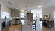 Located on Langdon Road in Morrisville, NC, this single-floor home offers a well-designed open-concept layout that seamlessly connects the kitchen, dining, and living areas. The kitchen is a standout feature, showcasing granite countertops, gray shaker-style cabinetry, and a central island with bar seating — ideal for both meal prep and casual dining. Pendant lighting hangs above the island, complementing the track lighting and flush-mount fixtures that illuminate the space. Stainless steel appliances, including a dishwasher, add a modern, functional touch. A window with a decorative Roman shade brings in natural light over the sink area. The kitchen flows effortlessly into the living room, which is brightened by large windows and anchored by a ceiling fan. The living space is open and inviting, with warm hardwood flooring that extends throughout the main areas, tying the entire floor plan together cohesively. A built-in shelving unit provides additional storage and display space between the kitchen and living zones. The overall ambiance is clean, contemporary, and welcoming, with a neutral palette of whites and grays accented by natural wood tones — making this Morrisville home both stylish and practical.