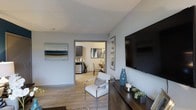 This single-floor residence offers a well-connected layout where the living area flows seamlessly into the dining and kitchen spaces. The living room is anchored by a large wall-mounted flat-screen television positioned above a dark wood media console, which is styled with decorative accents including a blue ceramic vase and small succulents. A modern accent chair with a chrome frame and neutral upholstery adds a sleek, contemporary touch to the seating area. Light wood-look flooring extends throughout, contributing to a bright and cohesive aesthetic. The walls are painted in a soft gray tone and adorned with framed artwork, reinforcing a calm, neutral palette. An open passageway connects the living area to the dining space, where a small dining table and chairs are visible, and natural light filters in from an adjacent window. The kitchen, visible in the background, features dark cabinetry and light countertops, offering a functional and stylish cooking space. The overall ambiance is modern and understated, with thoughtful décor choices that create a welcoming and well-appointed environment throughout.