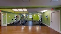 This single-floor fitness center offers a dedicated and fully equipped space for a comprehensive workout experience. The room is immediately distinguished by its bold, vibrant green walls, which create an energizing atmosphere conducive to exercise. The ceiling features recessed lighting panels that provide bright, even illumination throughout the space, complemented by an overhead ceiling fan to aid in air circulation during workouts.

The exercise area is outfitted with a variety of cardio equipment, including treadmills and elliptical trainers, arranged neatly along one side of the room. A multi-functional cable and strength training machine anchors the opposite side, offering a range of resistance exercises. Additional free-standing workout benches and open floor space provide room for stretching and bodyweight training.

Multiple wall-mounted flat-screen TVs are positioned at comfortable viewing angles, allowing users to stay entertained while exercising. The flooring transitions from rich hardwood near the entrance to heavy-duty rubber matting in the main workout area, providing both durability and safety underfoot. A window along the back wall allows for natural light to filter into the space. Overall, this fitness center is thoughtfully designed to support a well-rounded exercise routine in a motivating and functional environment.