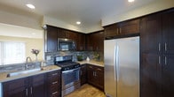 This single-floor space showcases a thoughtfully designed kitchen that serves as its centerpiece. The kitchen features rich, dark espresso-toned cabinetry that extends from floor to ceiling, providing generous storage capacity through both upper and lower cabinets with sleek bar-pull hardware. A striking mosaic tile backsplash in tones of gray and brown adds texture and visual interest to the walls, complementing the overall color palette beautifully. The kitchen is equipped with a full suite of stainless steel appliances, including a gas range with a built-in over-the-range microwave and a large side-by-side refrigerator, offering both functionality and a cohesive, contemporary aesthetic. Light-toned countertops provide a pleasing contrast against the dark cabinetry, creating a balanced and inviting workspace. Warm hardwood flooring grounds the space, adding a natural element that softens the modern design. Recessed lighting in the ceiling ensures the kitchen is well-lit, enhancing the clean lines and finishes throughout. Small decorative touches, including potted greenery on the countertops, contribute to the space's welcoming and lived-in ambiance.