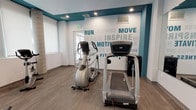 This fitness center is designed to provide a comprehensive workout environment within a compact, efficiently arranged space. The room is finished with durable wood-style flooring and features a crisp white and teal color scheme, with the teal accent prominently displayed along the upper walls and trim. A standout design element is the large motivational word-art mural covering one wall, featuring phrases such as "Run," "Inspire," "Motivate," and "Fitness," lending an energetic and encouraging atmosphere to the space.

The facility is equipped with a well-rounded selection of cardio machines, including treadmills, elliptical trainers, an upright stationary bike, and a cable machine. A dumbbell rack with a full range of free weights is positioned against the mirrored wall, which spans a significant portion of the room and visually expands the space. A flat-screen television is mounted above the dumbbell station, and an adjustable workout bench adds versatility to the strength training area.

Natural light filters into the room through multiple windows fitted with roller shades, complemented by recessed ceiling lighting. A structural column near the window wall adds an architectural element to the room's character. The overall layout is thoughtfully organized to maximize usability, making this fitness center a functional and motivating space for daily exercise.