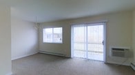 This single-floor residence offers a comfortable and functional layout filled with natural light. The main living and dining area serves as the centerpiece of the space, characterized by soft neutral-toned walls and wall-to-wall carpeting that creates a warm, inviting atmosphere. A sliding glass door with vertical blinds opens directly to an outdoor area, allowing abundant natural light to pour into the room while providing convenient outdoor access. A standard window with horizontal blinds further enhances the brightness of the space. An elegant chandelier adds a refined decorative touch overhead, lending a sense of classic style to the room. A baseboard heating system runs along the exterior wall, ensuring year-round comfort. The overall design is clean and understated, with a neutral color palette that offers a versatile backdrop for a variety of interior styles. The open flow of the main living area provides a sense of spaciousness and ease of movement throughout the home.